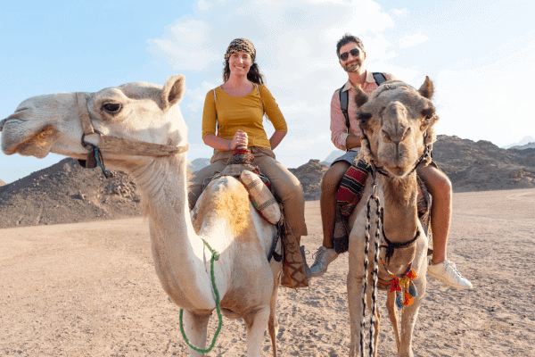 Two people riding camels in a desert landscape.