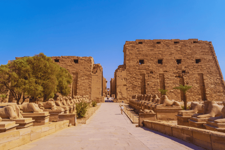 Pathway lined with sphinx statues and ancient ruins.